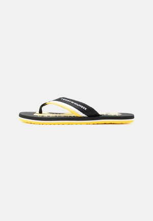 Tongs Tommy Hilfiger noires et jaunes avec brides d'orteils en tissu et nom de la marque sur la bride et la semelle, présentées de profil sur fond blanc.