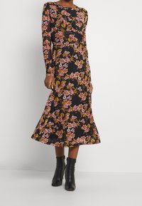 Svart blommig klänning med långa ärmar, prydd med rosa och gula blommor. Midilängd med en utsvängd kjol och bärs med svarta ankelstövlar.