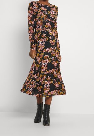 Robe noire à fleurs avec manches longues, ornée de fleurs roses et jaunes. Longueur midi avec une jupe évasée et portée avec des bottines noires.