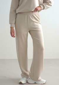 Beige, hochgeschnittene Sweatpants mit entspannter Passform und elastischem Bund, aus weichem Stoff gefertigt. Kombiniert mit weißen Sportschuhen.