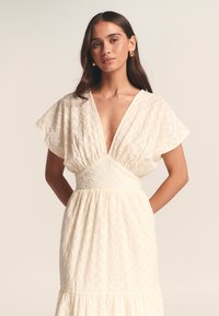 Hvid kjole med mønster, dyb V-udskæring, flagrende ærmer og en figursyet talje. Tekstureret stof med et flydende, lagdelt skørt.