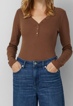 Vrouw draagt een bruin, geribd henley-shirt met lange mouwen en drie knopen, gecombineerd met een hoge taille blauwe spijkerbroek, handen in de zakken.