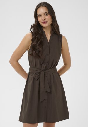 Femme aux longs cheveux foncés portant une robe sans manches marron foncé boutonnée et nouée à la taille, debout, les mains dans le dos.