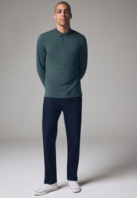 Langærmet mørkegrøn polo med krave, parret med mørkeblå jeans og hvide sneakers, stående mod en neutral baggrund.