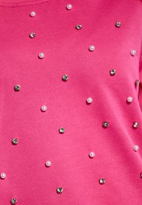 Maglione rosa con accentuazioni decorative rotonde: borchie silver lucide e piccole perle bianche disposte lungo il tessuto. Texture liscia.