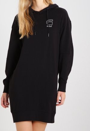 Kvinde iført sort langærmet hoodie-kjole med lille hvidt logo på brystet, hænderne ved siderne, stående mod en ensartet lys baggrund.