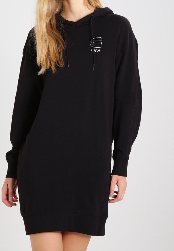 Femme portant une robe à capuche noire à manches longues avec un petit logo blanc sur la poitrine, mains près des côtés, debout contre un fond clair uni.