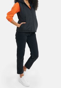 Zwarte gewatteerde bodywarmer met capuchon en ritssluiting, zijzakken, gecombineerd met een oranje trui en zwarte jeans, afgewerkt met witte sneakers.