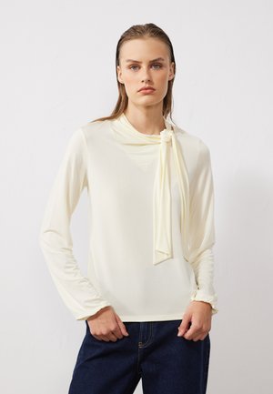 Cream long-sleeve top with a draped neckline and tie detail, featuring gathered sleeves and a smooth, soft texture. Worn with dark denim jeans.
