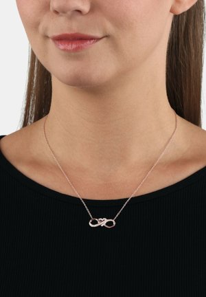 Frau, die eine zarte Halskette mit einem roségoldenen Unendlichkeit-Anhänger und kleinen eingearbeiteten Kristallen an einer dünnen Kette über einem schwarzen Oberteil trägt.