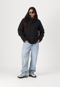 Svart vanntett jakke med høy krage, avslappet passform, kombinert med lyseblå baggy jeans og grå sportssko. Inkluderer solbriller.