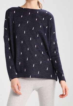 Maglietta a maniche lunghe blu realizzata in tessuto morbido con motivi di fulmine bianchi su tutto il tessuto. Vestibilità rilassata con scollatura ampia, abbinata a pantaloni grigio chiaro.