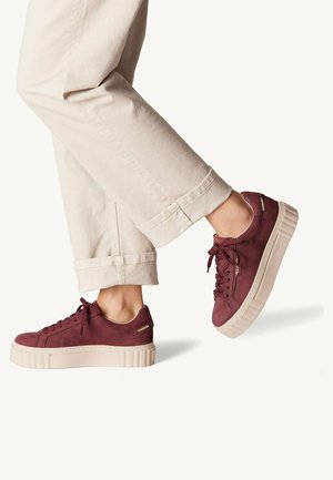 Personne portant un pantalon beige et des baskets à plateforme grenat, levant légèrement un pied sur un fond clair uni.