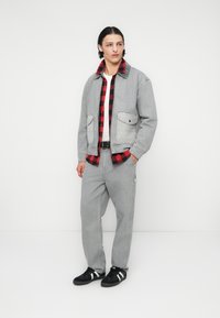 Jachetă din denim gri purtată peste o cămașă cu carouri roșii și negre, asortată cu pantaloni gri deschis și adidași negri cu dungi albe.