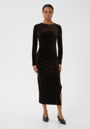 Femme portant une longue robe noire en velours à manches longues et fente latérale, associée à des chaussures noires à talons, debout devant un fond blanc.
