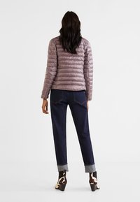 Kvinde med langt mørkt hår iført en mauve pufferjakke, mørke opfoldede jeans og sort-hvide hælboot, stående mod en hvid baggrund.