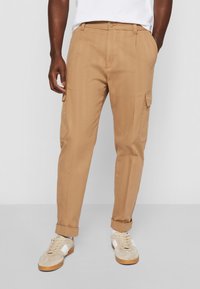 Beige Cargo-Hosen mit taillierter Passform, seitlichen Taschen, einem glatten Baumwollmaterial und umgeschlagenen Säumen, kombiniert mit hellen Sneakern.