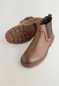 Braune Leder Chelsea-Stiefel mit elastischen Seitenpaneelen und robusten profilierten Sohlen, vor einem hellen Hintergrund präsentiert.