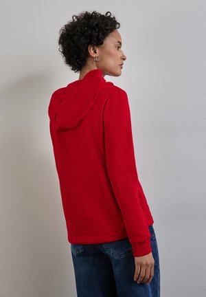 Sudadera roja con capucha, de mangas largas y puños acanalados, combinada con jeans azules. La tela parece suave con una textura lisa.