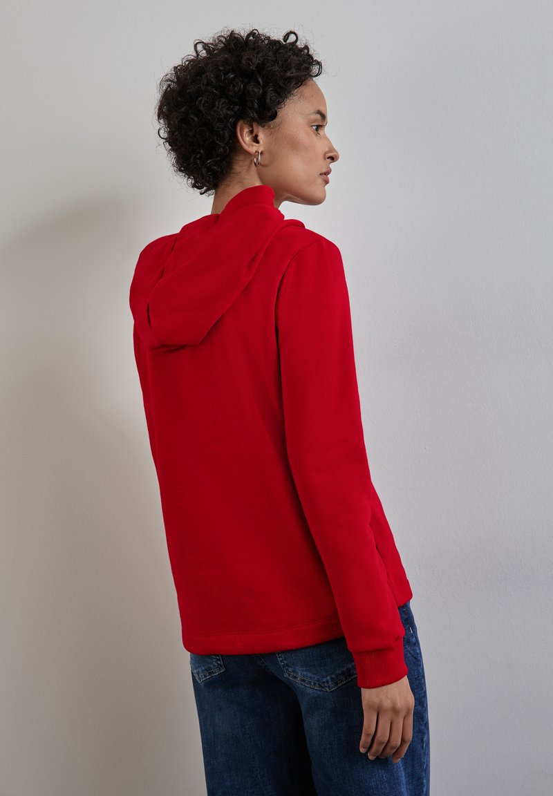 Felpa rossa con cappuccio, maniche lunghe e polsini a coste, abbinata a jeans blu. Il tessuto appare morbido con una superficie liscia.