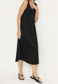 Svart ärmlös klänning i en avslappnad passform, tillverkad av ett glatt tyg, med knälång fåll och en enkel design. Bärs med platta sandaler.