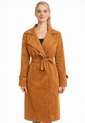 Blonde Frau trägt einen knielangen braunen Trenchcoat mit Gürtel, breiten Revers und geknöpften Manschetten über einem schwarzen Top.