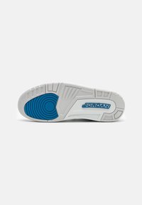 Semelle de basket blanche avec grip circulaire bleu et marquage "JORDAN" en bleu sur la zone de la voûte plantaire, présentée sur un fond blanc uni.
