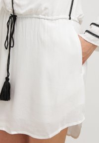 Gros plan sur une robe blanche avec un bord brodé noir et une ceinture à pompons noirs, montrant une main reposant dans une poche latérale.