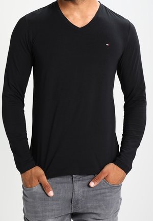 Chemise noire à manches longues en V, fabriquée en tissu doux, au design ajusté avec un petit logo sur la poitrine, associée à un jean gris clair.