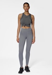 Grå kort atletisk linne knutet framtill, kombinerat med höga grå tights och svarta träningsskor. Enfärgad vit bakgrund.