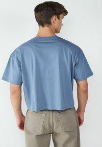 Hellblaues, kurzes Baumwoll-T-Shirt mit kurzen Ärmeln, rundem Ausschnitt und glatter Textur. Kombiniert mit sandfarbenen Hosen.