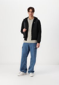 Črna zip-up jakna s kapuco, siva majica in modri široki kavbojki z stranskimi žepi. Model drži majhno črno torbico čez ramo.