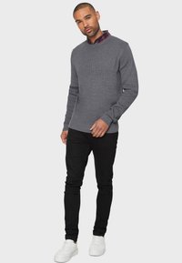 Maglione grigio lavorato a maglia con scollatura a girocollo, indossato sopra una camicia a quadri, abbinato a pantaloni neri slim-fit e sneakers bianche.