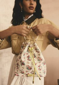 Blomster-mønstrede bucket bag med hvidt lærred, limegrøn snøring og gyldne hardware-detaljer, båret af en model i en beige skjorte.