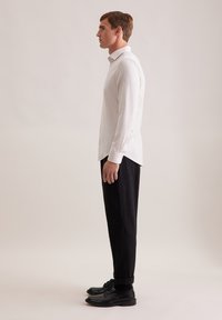 Camicia bianca a maniche lunghe con colletto button-down, tessuto testurizzato; abbinata a pantaloni neri slim e scarpe nere, vista di lato.