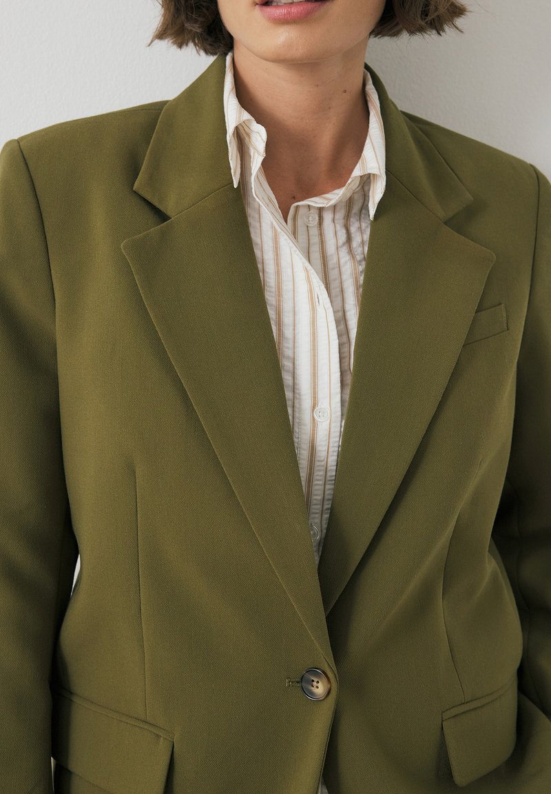 Blazer verde oliva con design strutturato, chiusura con un bottone, revers a punta e taschino applicato, indossato sopra una camicia bianca a righe.