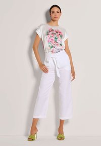 Witte linnen wijde broek gecombineerd met een kortemouwen, lichtkleurig top met een bloemenontwerp in roze en groen. Groene sandalen met banden.