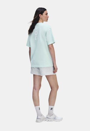 Mujer de perfil vistiendo una camiseta holgada azul claro, pantalones cortos grises, calcetines blancos y zapatillas deportivas blancas con marca negra.