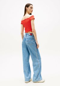 Top smock rosso a spalle scoperte abbinato a pantaloni larghi blu chiaro con una cucitura laterale bianca e un'etichetta del marchio ben visibile in vita.