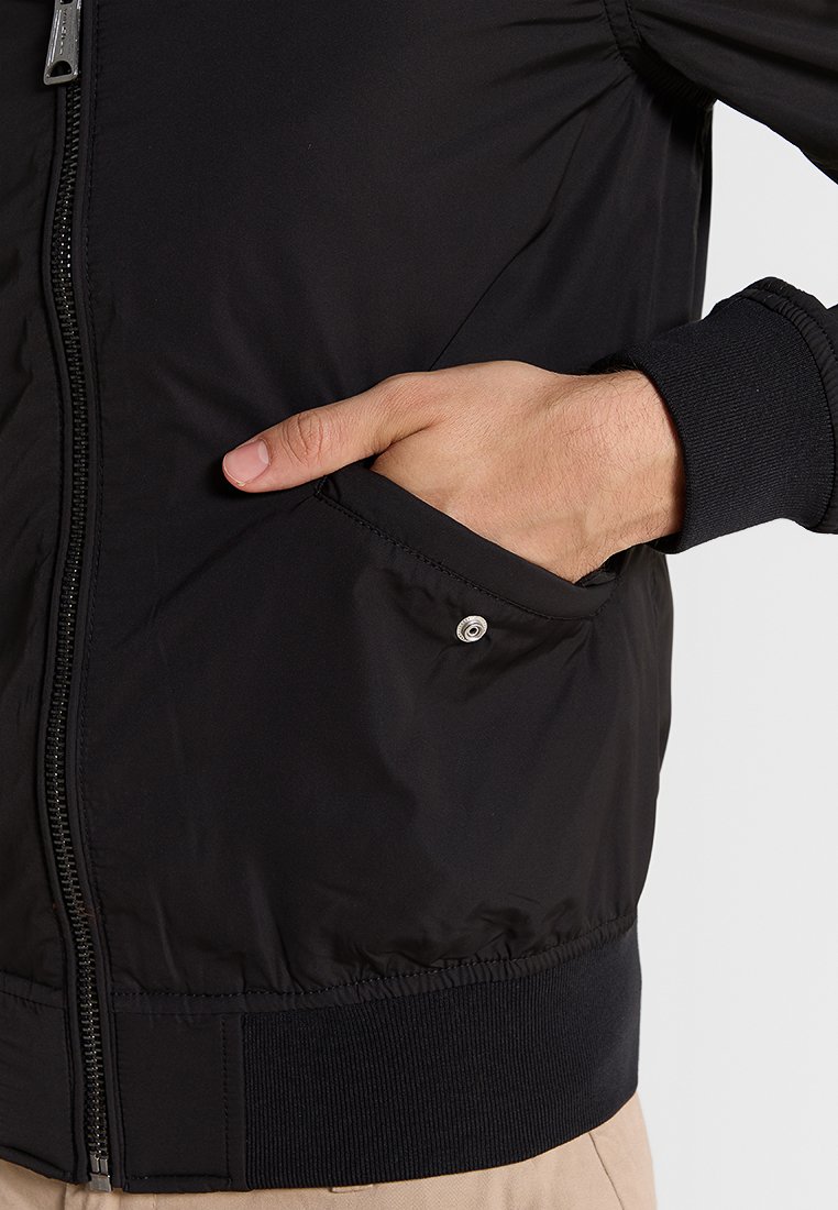 Veste bomber noire à fermeture éclair avec des poignets et un ourlet côtelés. Présente une poche latérale avec une fermeture à bouton-pression et un tissu lisse et léger.
