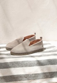 Beige Canvas-Slipper mit abgerundeter Zehe, Jutesohle und genähten Details. Auf gestreiftem Stoffhintergrund arrangiert.