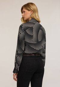 Vrouw met blond haar, gekleed in gouden ringoorbellen, zwarte broek en een zwart shirt met lange mouwen met een witte hartstip patroon, zijwaarts gekeerd.