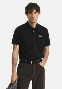 Man met krullend haar, gekleed in een zwart GANT polo shirt, donkere broek, zwarte riem, met arm tattoos en houdt een gestructureerde doek vast.