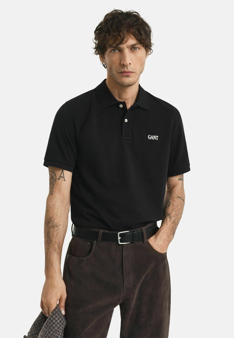 Man met krullend haar, gekleed in een zwart GANT polo shirt, donkere broek, zwarte riem, met arm tattoos en houdt een gestructureerde doek vast.