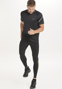 Musta urheilupaita, jossa on heijastavat yksityiskohdat, yhdistettynä tiukkoihin mustiin legginsseihin, joissa on keltainen raita. Yhdistetty mustiin urheilukenkiin.