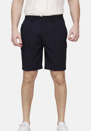 Uomo con pantaloncini neri su misura, camicia bianca e sneakers bianche, in piedi davanti a uno sfondo semplice.