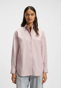 Light pink button-up shirt with a collar, featuring a curved hem and visible seams. Long sleeves with buttoned cuffs. Smooth texture.