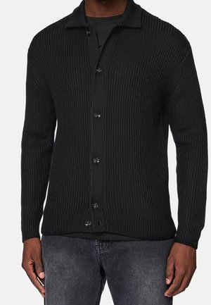 Cardigan noir côtelé à boutons porté avec un jean gris foncé et un T-shirt noir en dessous, visible des épaules aux hanches.