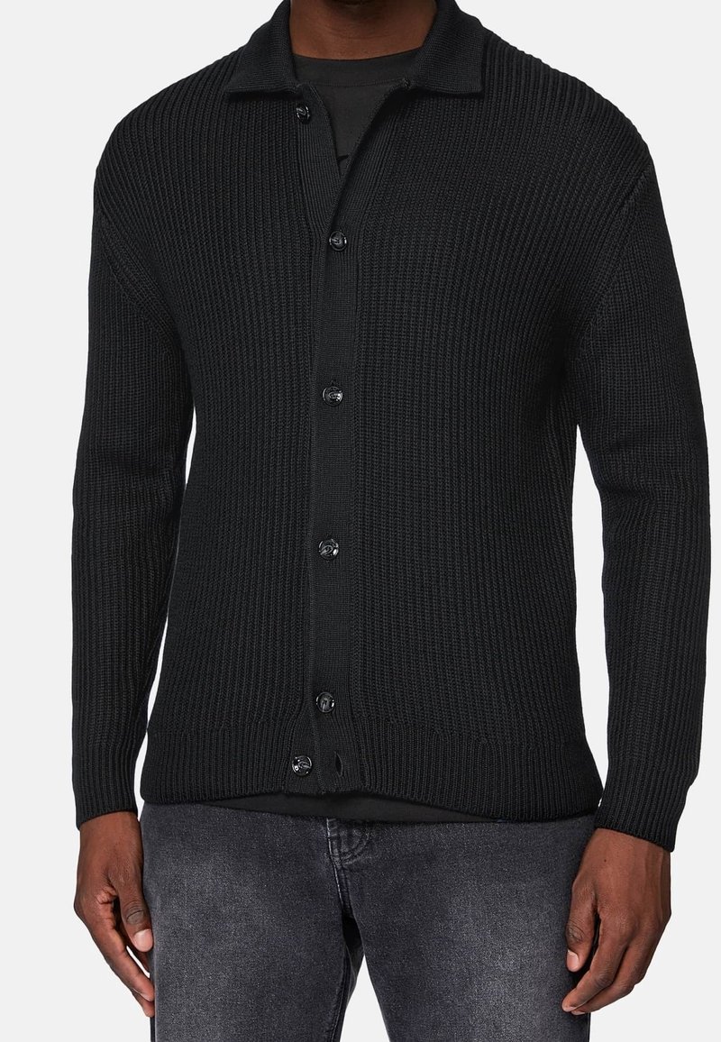 Cardigan noir côtelé à boutons porté avec un jean gris foncé et un T-shirt noir en dessous, visible des épaules aux hanches.
