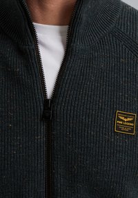 Donkergroene geribbelde zip-up trui met een hoge kraag, een bruine rits en een gele geborduurde logo patch op de borst.